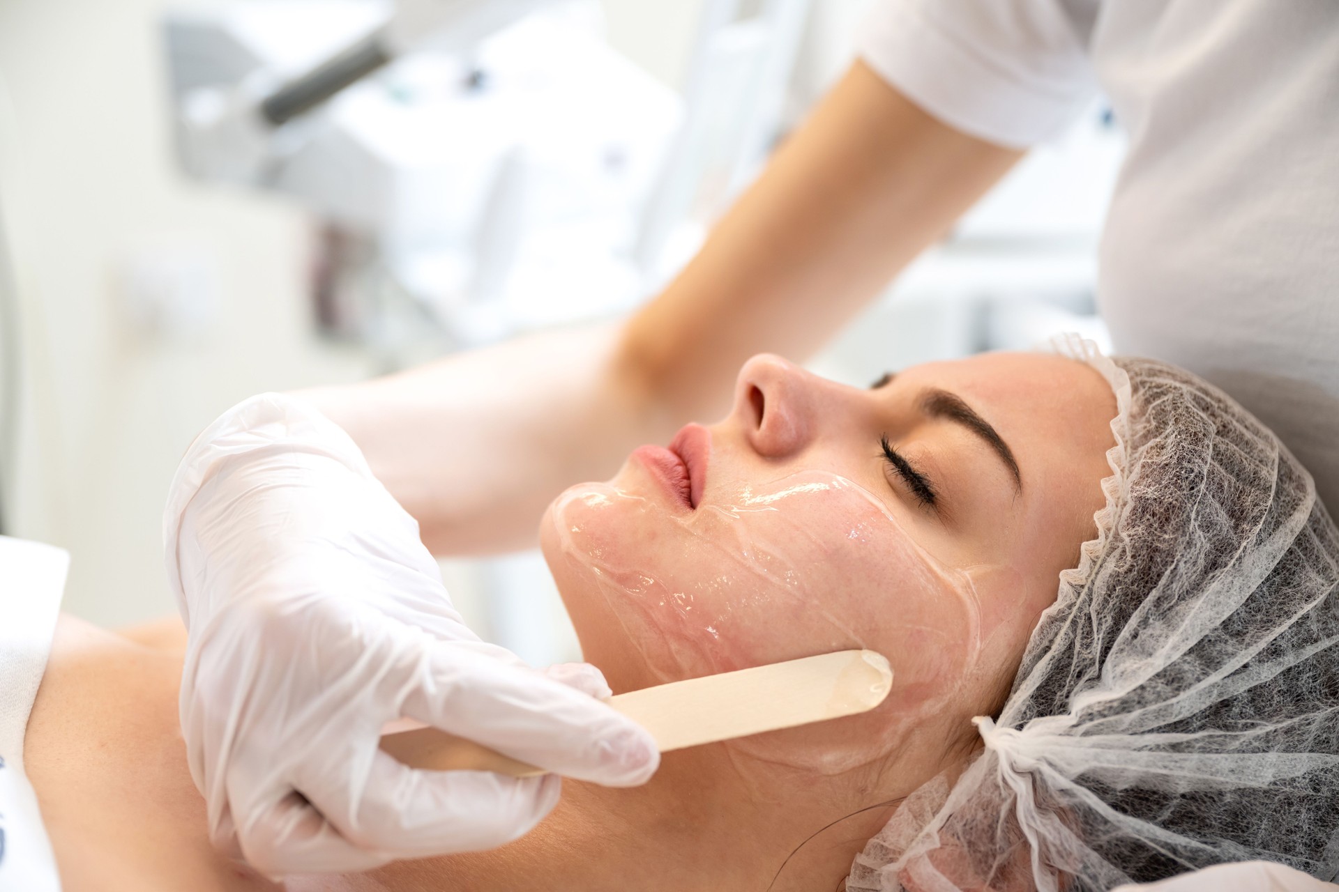 Closeup of beautician hands with wooden spatula applying cosmetic product gel peeling mask to beautiful young woman face