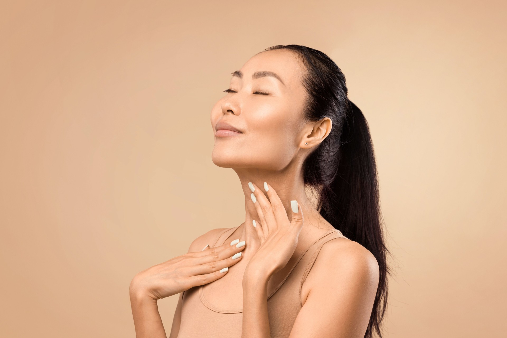 Middle-aged Asian woman touching smooth skin on neck, beige studio backdrop